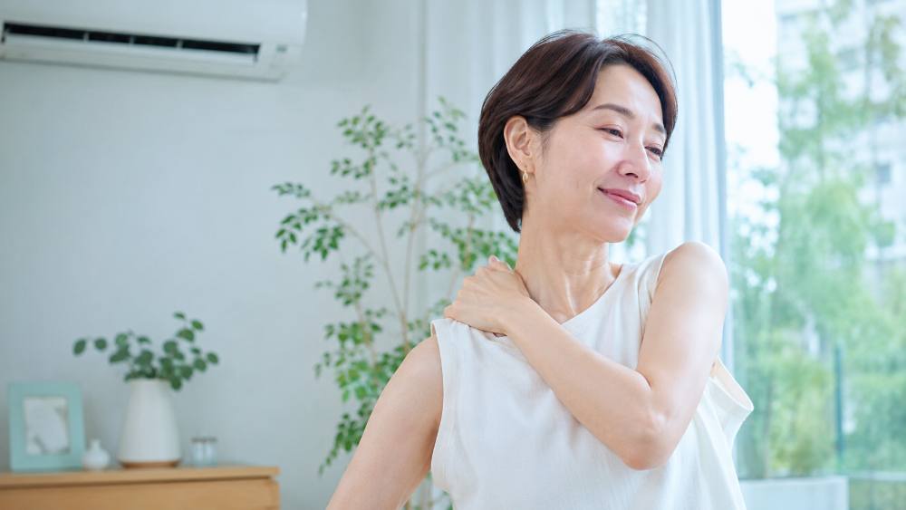 エアコンのある室内で肩に手を当てて肩をほぐす女性|夏の冷房と肩こり
